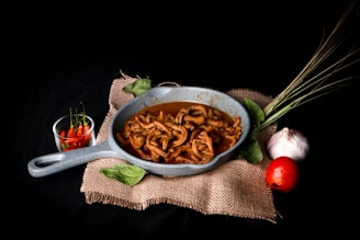 A rustic-looking grey skillet filled with cooked food, likely meat, resting on a piece of burlap cloth. Surrounding the skillet are two green leaves, a bunch of lemongrass, a white garlic bulb, and a red tomato. To the left of the skillet, there is a small glass bowl containing red chili peppers.