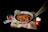 A rustic-looking grey skillet filled with cooked food, likely meat, resting on a piece of burlap cloth. Surrounding the skillet are two green leaves, a bunch of lemongrass, a white garlic bulb, and a red tomato. To the left of the skillet, there is a small glass bowl containing red chili peppers.