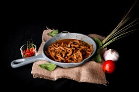 A rustic-looking grey skillet filled with cooked food, likely meat, resting on a piece of burlap cloth. Surrounding the skillet are two green leaves, a bunch of lemongrass, a white garlic bulb, and a red tomato. To the left of the skillet, there is a small glass bowl containing red chili peppers.