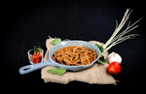 A rustic presentation of a dish featuring spiral-shaped cooked ingredients in a blue-gray pan. Accompanying the dish are fresh vegetables including a bunch of lemongrass, a whole garlic bulb, a red tomato, and a small bowl of red and green chili peppers, all arranged on a piece of burlap fabric against a dark background.