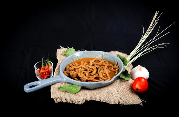 A rustic presentation of a dish featuring spiral-shaped cooked ingredients in a blue-gray pan. Accompanying the dish are fresh vegetables including a bunch of lemongrass, a whole garlic bulb, a red tomato, and a small bowl of red and green chili peppers, all arranged on a piece of burlap fabric against a dark background.