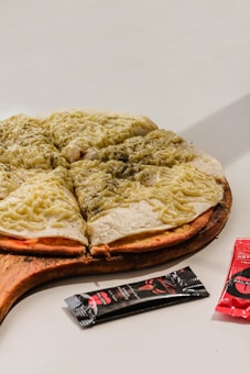 A pizza topped with melted cheese and herbs is placed on a wooden serving board. The pizza is cut into several slices. Next to the pizza, there are packets of condiments, likely sauce, with black and red packaging.