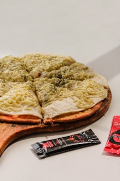 A pizza topped with melted cheese and herbs is placed on a wooden serving board. The pizza is cut into several slices. Next to the pizza, there are packets of condiments, likely sauce, with black and red packaging.