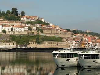 A riverside scene featuring two modern cruise boats docked in the foreground with scenic reflections on the water. In the background, there is a hillside populated with a mix of colorful buildings and lush green trees. The architecture appears traditional, with some buildings having red tile roofs, and the overall environment is calm and picturesque.