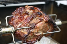 A large, juicy piece of meat is roasting on a spit, with a beautifully browned and crispy exterior. The roast is being cooked using a rotisserie method, evident from the metal skewer piercing through it, rotating over a grill.