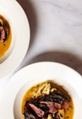 Two white plates with gourmet-style cooked meat and grilled vegetables, presented with a rich sauce on a light-colored marble surface. One plate is positioned prominently in the foreground, while the edge of another plate is visible at the top left corner.