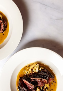 Two white plates with gourmet-style cooked meat and grilled vegetables, presented with a rich sauce on a light-colored marble surface. One plate is positioned prominently in the foreground, while the edge of another plate is visible at the top left corner.