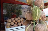A market stall features several durians, some in a large pile within a white crate and others hanging on a structure to the right. The background includes signage with text and a mural depicting plants. A person in red clothing and a green cap stands behind the crate.