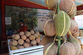 A market stall features several durians, some in a large pile within a white crate and others hanging on a structure to the right. The background includes signage with text and a mural depicting plants. A person in red clothing and a green cap stands behind the crate.