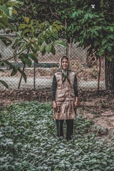 An elderly person stands in a garden surrounded by green plants and foliage. The background features a metal fence and a variety of lush trees. The person is wearing a headscarf and a long vest-like coat, with a calm, contemplative expression.