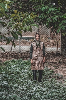 An elderly person stands in a garden surrounded by green plants and foliage. The background features a metal fence and a variety of lush trees. The person is wearing a headscarf and a long vest-like coat, with a calm, contemplative expression.