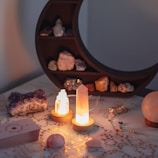 A collection of various crystals and stones displayed on a surface with a crescent moon-shaped shelf in the background. The scene is softly lit, highlighting the translucent quality of the crystals, which are placed on small wooden stands. Decorative stars are scattered across the marble surface, alongside a deck of cards and a pendulum with colorful beads.
