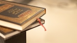 Two books with dark, intricately designed covers and golden embossed text are stacked, with a red ribbon bookmark peeking out.