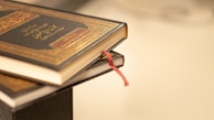 Two books with dark, intricately designed covers and golden embossed text are stacked, with a red ribbon bookmark peeking out.