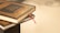 Two books with dark, intricately designed covers and golden embossed text are stacked, with a red ribbon bookmark peeking out.