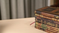 A stack of ornately designed books with Arabic script on their spines lies on a light wooden surface. The books feature intricate gold and red details and are neatly stacked in descending order. In the background, muted curtains add a soft, elegant touch.