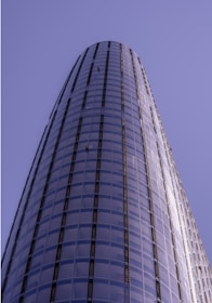 A tall, cylindrical skyscraper with a sleek glass facade extends upwards into a clear blue sky. Two window cleaners in red uniforms are visible on the exterior, highlighting the building's impressive height and modern architecture.
