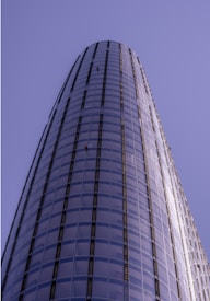 A tall, cylindrical skyscraper with a sleek glass facade extends upwards into a clear blue sky. Two window cleaners in red uniforms are visible on the exterior, highlighting the building's impressive height and modern architecture.