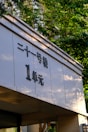 A building facade featuring Chinese characters prominently displayed on a white surface. Sunlight filters through the green leaves of nearby trees, casting subtle shadows on the structure.