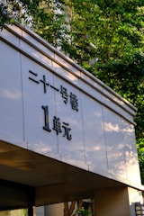 A building facade featuring Chinese characters prominently displayed on a white surface. Sunlight filters through the green leaves of nearby trees, casting subtle shadows on the structure.