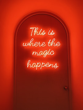 A neon sign with the phrase 'This is where the magic happens' glows with a warm, orange hue filling a curved doorway. The lighting creates a cozy and mystical ambiance.
