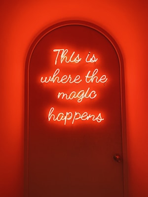A neon sign with the phrase 'This is where the magic happens' glows with a warm, orange hue filling a curved doorway. The lighting creates a cozy and mystical ambiance.