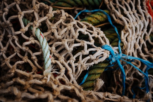 A tangled arrangement of ropes and nets in various colors, primarily beige and blue. The texture of the nets is prominent, showcasing intricately woven patterns. Some ropes are multicolored, including light green and red tints. The overall appearance is rugged and maritime.