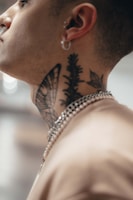 A close-up side profile of a person with intricate tattoos on their neck, including designs of a butterfly, leaves, and plants. The individual is wearing a chain neck accessory and has a small hoop earring in their ear.