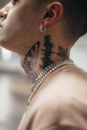 A close-up side profile of a person with intricate tattoos on their neck, including designs of a butterfly, leaves, and plants. The individual is wearing a chain neck accessory and has a small hoop earring in their ear.