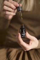 A pair of hands holding a small dark bottle labeled 'H Drop Joys of Spring Full Spectrum Hemp Oil' with a dropper in the other hand, releasing oil back into the bottle. The person is wearing a brown outfit, and the background is blurred.
