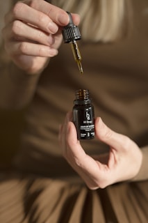 A pair of hands holding a small dark bottle labeled 'H Drop Joys of Spring Full Spectrum Hemp Oil' with a dropper in the other hand, releasing oil back into the bottle. The person is wearing a brown outfit, and the background is blurred.