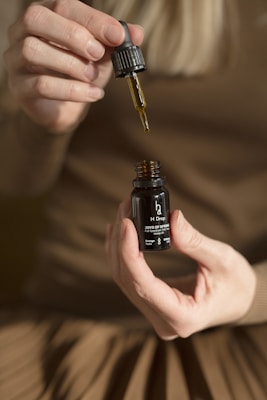 A pair of hands holding a small dark bottle labeled 'H Drop Joys of Spring Full Spectrum Hemp Oil' with a dropper in the other hand, releasing oil back into the bottle. The person is wearing a brown outfit, and the background is blurred.
