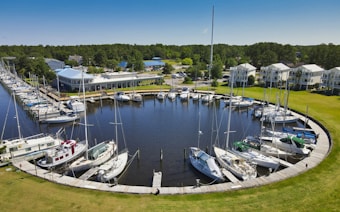 A serene marina with numerous sailboats and yachts docked in a circular arrangement. Surrounding the marina, there are green lawns and several multi-story residential buildings. A spacious clubhouse with a distinctive blue roof sits on one side. The scene is bathed in bright sunlight under a clear blue sky.