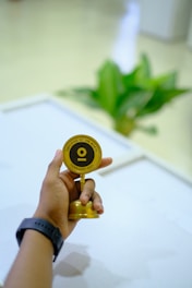 A hand holding a small golden trophy labeled 'Employee of the Month February 2021' with a blurred background featuring a green potted plant.