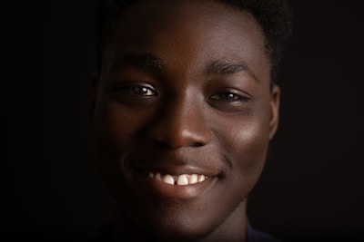 A close-up portrait of a person with a warm smile, set against a dark background. The lighting highlights their facial features, creating a gentle contrast.