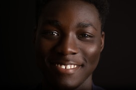 A close-up portrait of a person with a warm smile, set against a dark background. The lighting highlights their facial features, creating a gentle contrast.