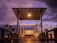 An outdoor deck with a wooden swing under a pergola, featuring warm lighting from a lamp above. The sky displays a stunning sunset with purple and orange hues, and the deck is adorned with potted plants and lanterns that add a cozy ambiance.
