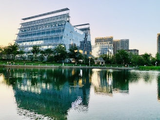 A modern commercial complex with large glass buildings is situated near a tranquil pond with reflections of the buildings in the water. There are trees and green spaces surrounding the buildings, and a few people are walking around the vicinity.