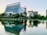 A modern commercial complex with large glass buildings is situated near a tranquil pond with reflections of the buildings in the water. There are trees and green spaces surrounding the buildings, and a few people are walking around the vicinity.