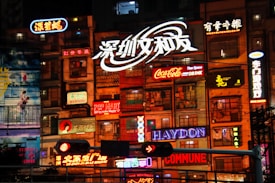 A vibrant urban scene featuring multiple colorful neon signs on the facade of a building at night. The signs display various brands, logos, and Chinese characters, illuminating the area with bright lights. The architecture of the building appears to be a mix of residential and commercial, with windows and balconies visible.