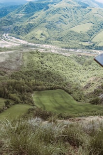 A scenic view of lush green hills and a well-maintained golf course surrounded by dense forest. The landscape in the background includes rolling hills covered with trees and some exposed rock formations. A road can be seen winding through the valleys.