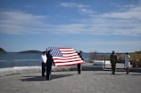 A group of people are standing outside by the water, holding a large American flag. There is a paved area in the foreground and a clear sky above. Some individuals are dressed in military uniforms while others are in civilian clothing.