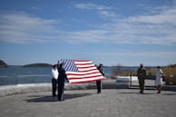 A group of people are standing outside by the water, holding a large American flag. There is a paved area in the foreground and a clear sky above. Some individuals are dressed in military uniforms while others are in civilian clothing.