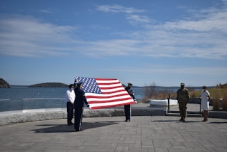 A group of people are standing outside by the water, holding a large American flag. There is a paved area in the foreground and a clear sky above. Some individuals are dressed in military uniforms while others are in civilian clothing.