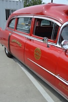 A vintage red ambulance with chrome detailing is parked outdoors next to a white container. The vehicle features the word 'AMBULANCE' and a fire department museum logo on the side. The windows are slightly tinted, and the interior appears to have old-fashioned seating.