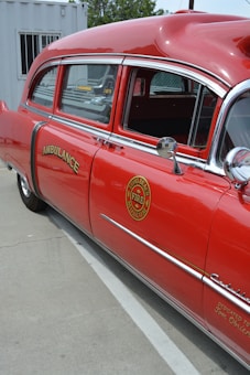 A vintage red ambulance with chrome detailing is parked outdoors next to a white container. The vehicle features the word 'AMBULANCE' and a fire department museum logo on the side. The windows are slightly tinted, and the interior appears to have old-fashioned seating.