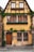An old, charming house with a timber-frame structure and yellow-painted walls. The building features multiple windows adorned with flower boxes full of greenery, climbing ivy, and a wooden door. The street in front is cobblestoned.