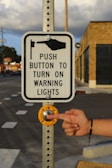 A metal pole holds a rectangular sign with a graphic and text instructing to push a button to activate warning lights. A hand is about to press the large, round, yellow button set into the pole. The background includes a building with brick walls and a road with painted crosswalk lines.