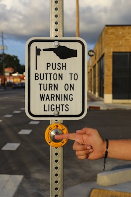 A metal pole holds a rectangular sign with a graphic and text instructing to push a button to activate warning lights. A hand is about to press the large, round, yellow button set into the pole. The background includes a building with brick walls and a road with painted crosswalk lines.