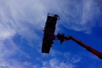 A large industrial crane is lifting a platform high into the sky, and a person is standing on the edge of the platform. The sky is partly cloudy with a mix of blue and white hues.
